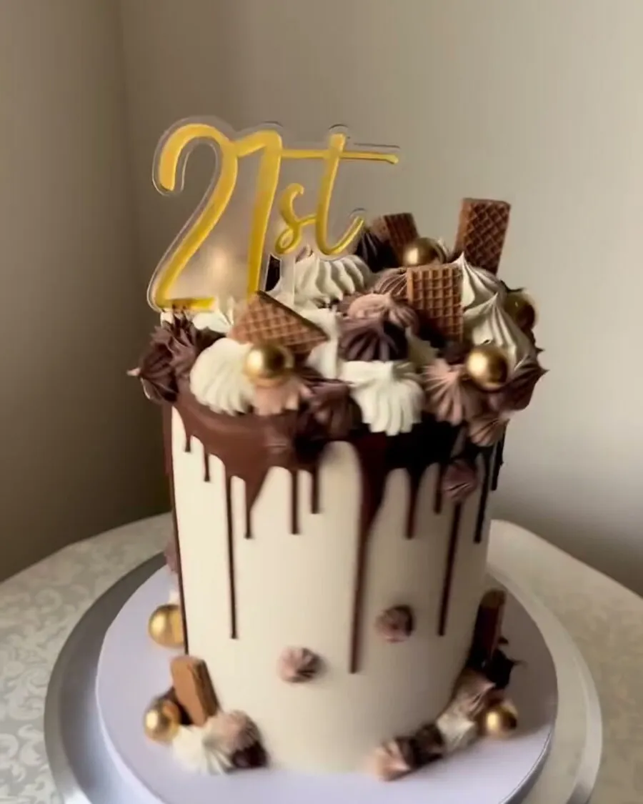 A tall cream buttercream cake with a chocolate drip, wafers, gold balls and a 21st topper