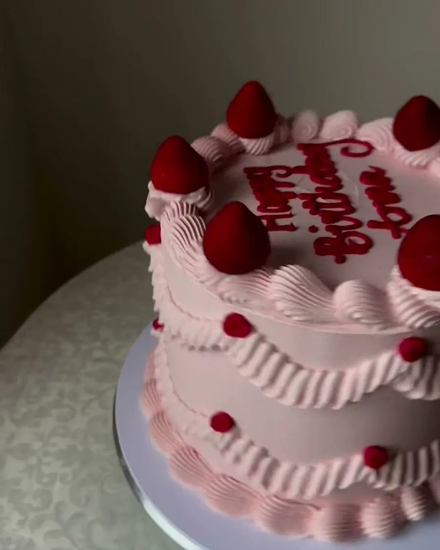 A pink buttercream cake with piped ruffles, red script lettering and fresh strawberries