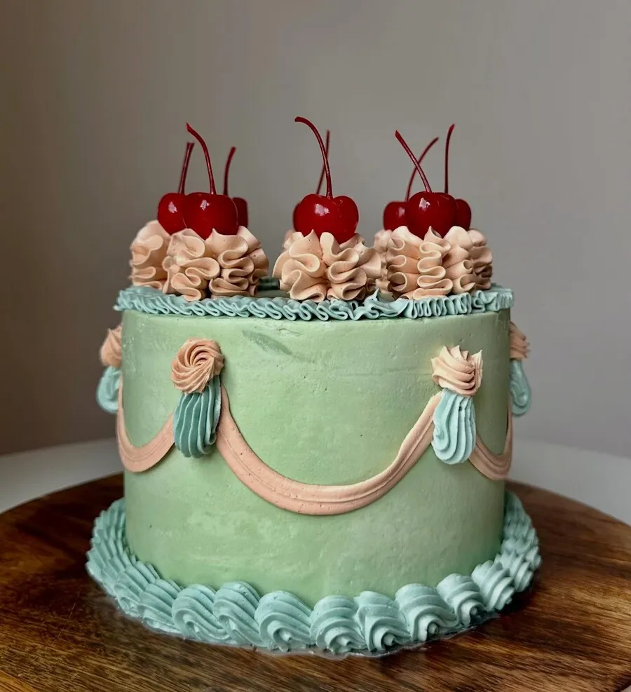 A sage green buttercream cake with peach piped swags, shell borders and glace cherries on top